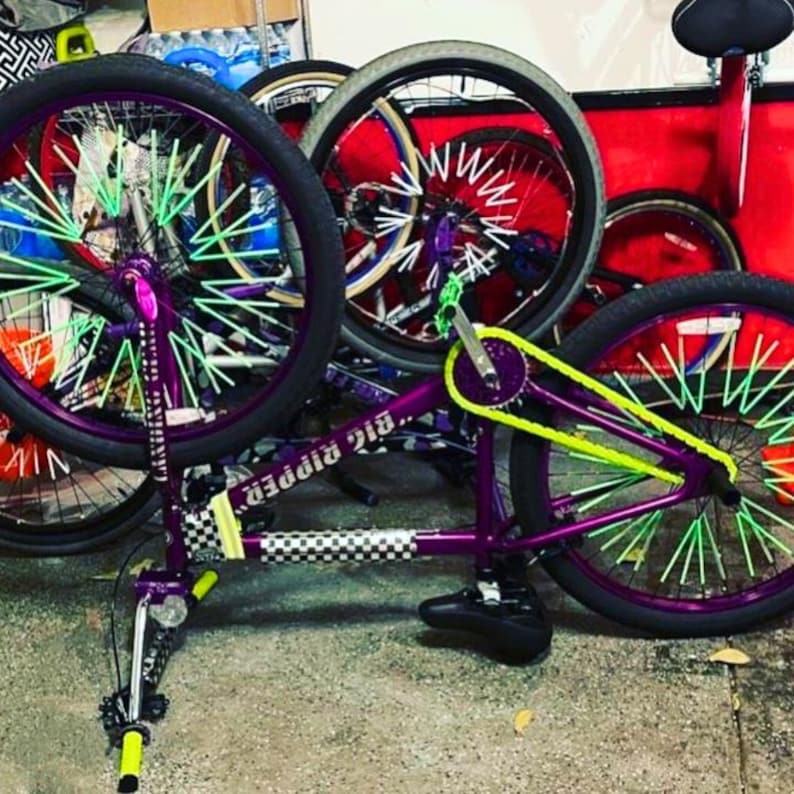 May include: A purple BMX bike with neon green and purple accents. The bike features a checkered pattern on the frame, neon green spokes, and a neon yellow chain. The wheels are black with purple rims.