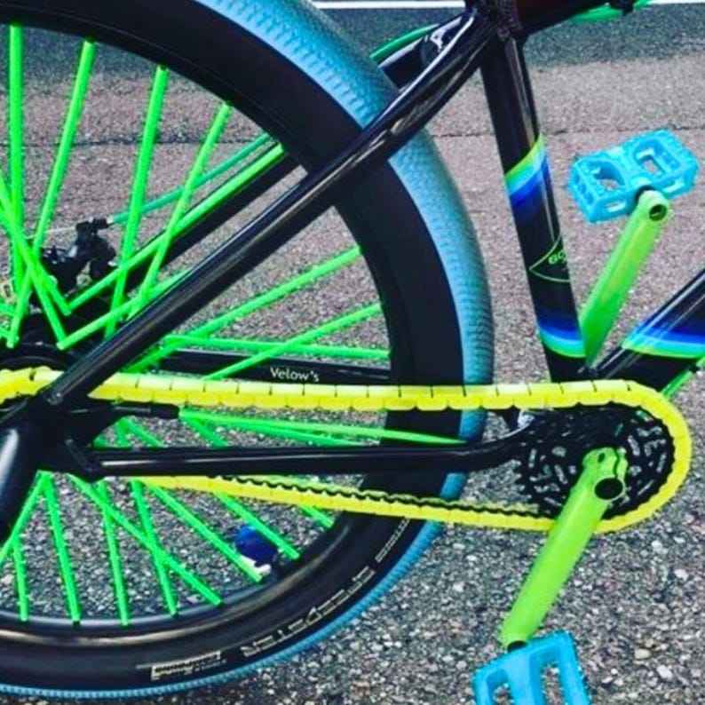 May include: Close-up of a custom bicycle wheel with neon green spokes and a black tire with a blue sidewall. The chain is yellow, and the pedals are blue. The frame is black with green and blue accents.