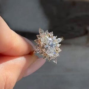 May include: A gold ring featuring a large, oval-cut diamond surrounded by smaller, sparkling stones. The ring's design resembles a sunburst, with the central diamond as the focal point. The ring is held up by a person's fingers.