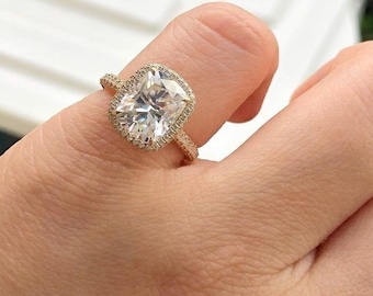 May include: A gold ring with a large, rectangular-cut, clear gemstone surrounded by a halo of smaller stones. The ring's band is also adorned with small stones. The ring is on a finger.