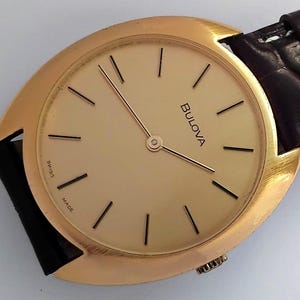 May include: A gold-tone Bulova wristwatch with a round, slightly oval-shaped face. The watch has a brown leather strap and a white dial with black hour markers and hands. The watch face says "Bulova" and "Swiss Made".