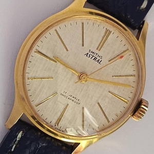 May include: A vintage gold-toned Smiths Astral wristwatch with a cream-colored face and gold hands. The watch features the text "SMITHS ASTRAL" and "17 JEWELS SHOCKPROOF". The watch has a dark blue leather strap.