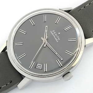May include: A Zenith automatic wristwatch with a grey face, silver hands, and hour markers. The watch has a date display and a grey leather strap. The words "ZENITH automatic" are printed on the watch face. The watch is marked "SWISS MADE".