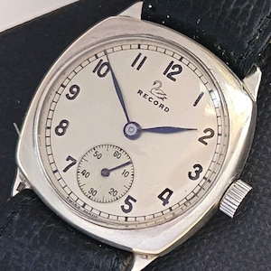 Op de afbeelding: Een vintage zilverkleurig polshorloge met een crèmekleurige wijzerplaat. Het horloge heeft zwarte cijfers, een kleine secondenwijzer en blauwe wijzers. De merknaam "RECORD" staat bovenaan in het midden. Zwarte leren band.