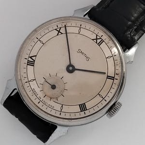 May include: A vintage Smiths wristwatch with a silver-colored face and black Roman numerals. The watch has a small seconds dial and a black leather band. The words "SMITHS" and "MADE IN ENGLAND" are visible on the watch face.