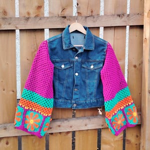May include: A denim jacket with crochet sleeves in magenta, turquoise, and orange. The sleeves have a floral pattern. The jacket is suspended on a wooden hanger against a wooden fence background.