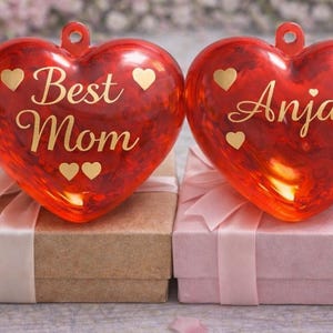 Könnte beinhalten: Zwei rote herzförmige Ornamente mit goldenem Text und Herzakzenten. Eines sagt "Best Mom", das andere "Anja". Sie liegen auf Geschenkboxen mit Schleifen. Im Hintergrund eine brennende Kerze.