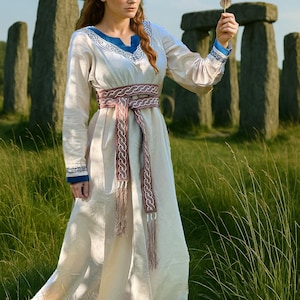May include: A cream-colored, long-sleeved dress with blue and white embroidered trim and a braided pink and brown belt. The dress is worn outdoors with a stone structure in the background. The person is holding a feather.