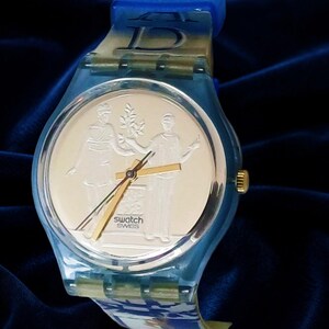 Puede incluir: Un reloj de pulsera Swatch con caja y correa de plástico azul translúcido. La esfera es de color dorado y presenta un diseño en relieve de dos figuras y una planta. Las palabras "swatch" y "SWISS" están impresas en negro en la esfera.