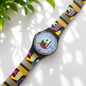 Könnte beinhalten: Eine Swatch-Armbanduhr mit rundem schwarzem Gehäuse und hellblauem Zifferblatt mit geometrischem Muster. Das Armband zeigt ein buntes abstraktes Design mit schwarzen, gelben, rosa und blauen Formen. Die Uhrzeiger sind dünn und metallisch.