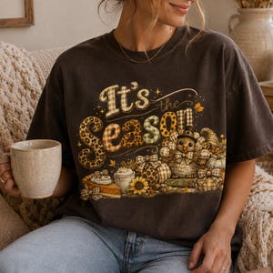 Puede incluir: Una camiseta de manga corta en color gris carbón oscuro con un estampado gráfico que incluye el texto "It's the Season" en una mezcla de patrones de leopardo y cuadros. El diseño muestra un pavo con sombrero, calabazas, tarta y girasoles.
