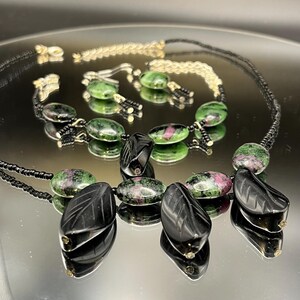 May include: A jewellery set comprising a necklace, bracelet, and earrings. The necklace and bracelet feature green and pink oval stones, black leaf-shaped pendants, and black beaded accents. The earrings have green oval stones. The set is displayed on a reflective surface.