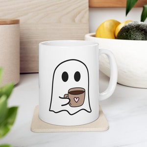 May include: White ceramic coffee mug with a ghost illustration holding a brown coffee cup with a pink heart. The ghost has two black eyes.