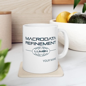 Può includere: Tazza in ceramica bianca con un design grafico in bianco e nero. Il design presenta il testo "Macrodatal Refinement" con un globo e la parola "Lumon" al suo interno. Il testo "Your Name" è in fondo al design.