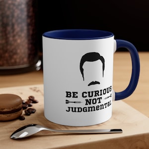 May include: White ceramic coffee mug with a blue rim and handle. The mug features a black silhouette of a man with a mustache and the text "Be Curious Not Judgmental" with a dart graphic.