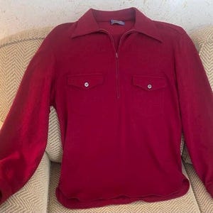 May include: A long-sleeved, burgundy-colored shirt with a collared neckline and a quarter-length zipper. The shirt features two chest pockets with button closures and a rounded hem. The fabric appears to be a soft, textured material.