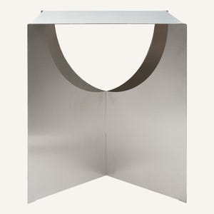 May include: A contemporary, silver side table with a distinctive geometric form. It has a rectangular top and a curved cutout on the front, forming an open, semi-circular space. The table is approximately 46 cm high and 38 cm wide.