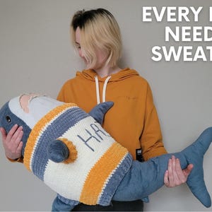 K&ouml;nnte beinhalten: Eine Person h&auml;lt ein blaues und wei&szlig;es geh&auml;keltes Hai-Pl&uuml;schtier mit dem Wort "Happy" bestickt darauf. Der Text "Every Haj needs a sweater!" ist in Wei&szlig; auf wei&szlig;em Hintergrund geschrieben.