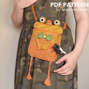 May include: An orange crochet crossbody bag shaped like a frog with large eyes, a green bow tie, and dangling legs. A small grey crocheted fly is attached to the side. The text PDF PATTERN by knotsinmybrain is visible in the upper right corner.