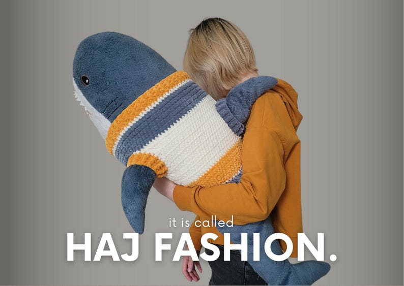 K&ouml;nnte beinhalten: Eine Person h&auml;lt einen gro&szlig;en blauen Pl&uuml;schhai, der einen gestreiften Pullover tr&auml;gt. Der Text "it is called HAJ FASHION." befindet sich unter dem Hai.