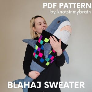 Könnte beinhalten: Ein Plüschhai mit einem bunten Pullover. Der Pullover ist schwarz mit einem Schachbrettmuster aus neonrosa, grünen, blauen und lila Quadraten. Der Text auf dem Bild lautet "PDF PATTERN by knotsinmybrain" und "BLAHAJ SWEATER."