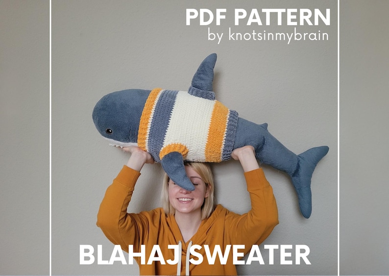 K&ouml;nnte beinhalten: Eine Person in einem gelben Pullover h&auml;lt ein blaues und wei&szlig;es geh&auml;keltes Hai-Spielzeug &uuml;ber ihrem Kopf. Der Hai tr&auml;gt einen gestreiften Pullover in Gelb, Wei&szlig; und Blau. Der Text "BLAHAJ SWEATER" ist unter dem Bild gedruckt. Der Text "PDF PATTERN by knots in my brain" ist &uuml;ber dem Bild gedruckt.