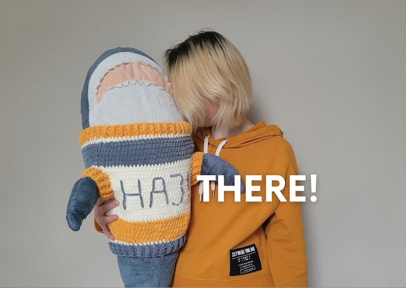 K&ouml;nnte beinhalten: Eine Person in einem gelben Hoodie mit dem Text "THERE!" h&auml;lt ein geh&auml;keltes Hai-Pl&uuml;schtier mit dem Text "HA3" auf der Vorderseite.