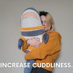 K&ouml;nnte beinhalten: Eine Person umarmt ein gro&szlig;es, graues und wei&szlig;es Pl&uuml;sch-Hai-Spielzeug, das einen gestreiften Pullover in Gelb, Grau und Wei&szlig; tr&auml;gt. Der Text "INCREASE CUDDLINESS." befindet sich am unteren Rand des Bildes.