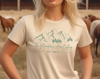 以下が含まれることがあります： 緑色の山脈のグラフィックと「The Mountains Are Calling and I Must Go」というテキストが書かれたベージュのTシャツを着た女性です。