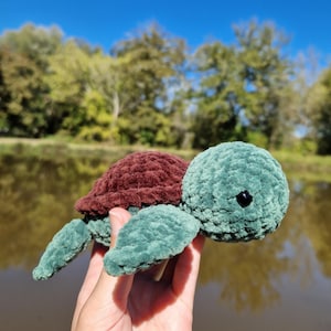 Peut inclure: Une tortue au crochet faite à la main avec une tête et des nageoires turquoise et une carapace bordeaux. Le jouet en peluche est présenté sur fond de lac et d'arbres sous un ciel bleu vif. La tortue a un œil noir.