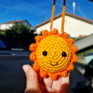 Op de afbeelding: Een gehaakte zonvormige versiering met een lachend gezicht en oranje stralen. De zon heeft zwarte ogen en een kleine, gebogen mond. De versiering wordt opgehangen door twee oranje koorden.