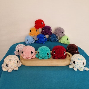 Puede incluir: Una colección de 14 peluches de pulpo tejidos a crochet en varios colores, incluyendo rojo, azul, verde, morado y naranja. Los peluches están dispuestos en forma de pirámide sobre una superficie azul.