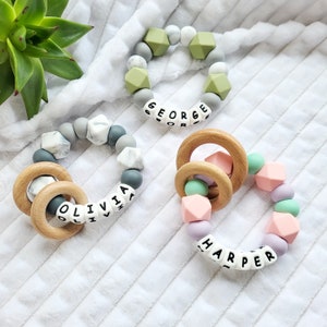 May include: Three silicone teething rings with wooden rings. The rings are in different colors and have the names "Olivia", "George", and "Harper" spelled out on them.