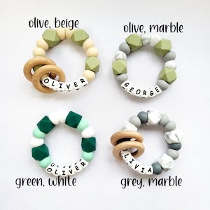 May include: Four silicone teething rings with wooden rings and personalized names. The rings are in different color combinations: olive and beige, olive and marble, green and white, and grey and marble.