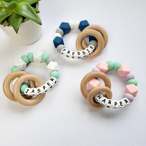 May include: Three silicone teething rings with wooden rings. The rings are in different colors and have the names "Freddie", "Jasper", and "Harper" spelled out on them.