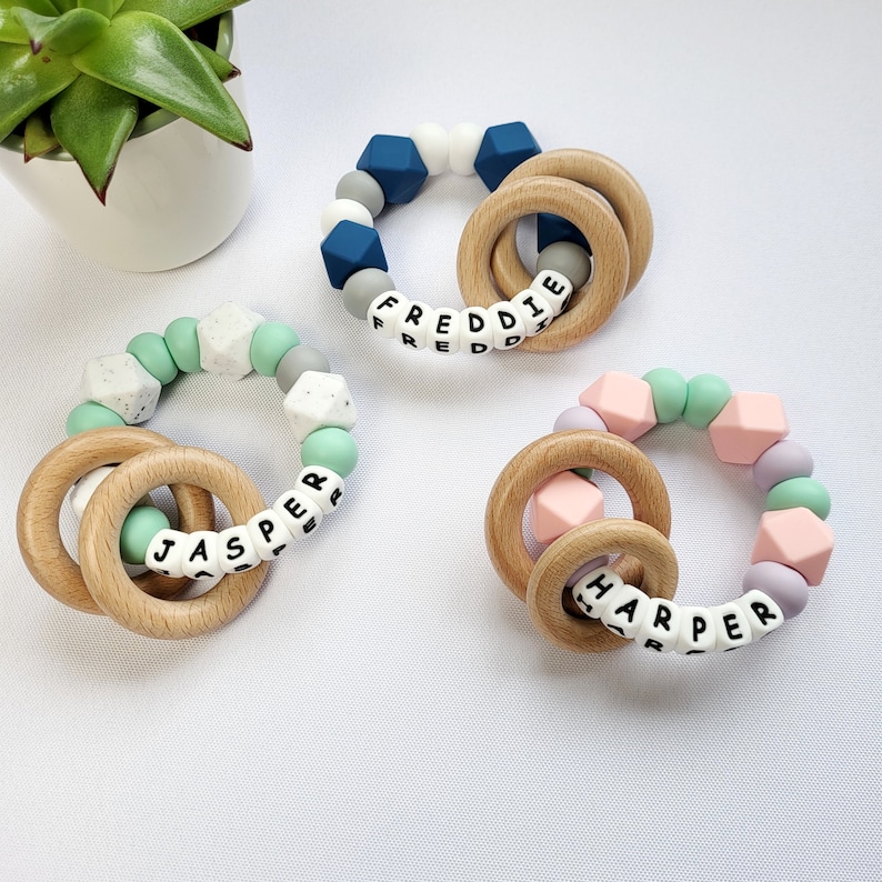 May include: Three silicone teething rings with wooden rings. The rings are in different colors and have the names "Freddie", "Jasper", and "Harper" spelled out on them.