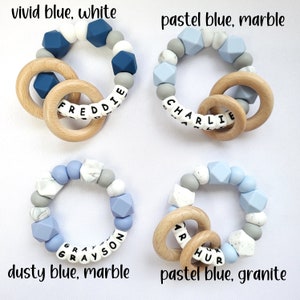 May include: Four silicone teething rings with wooden rings. The rings are in different colors and patterns: vivid blue and white, pastel blue and marble, dusty blue and marble, and pastel blue and granite. Each ring has a name spelled out in black letters: Freddie, Charlie, Grayson, and Arthur.