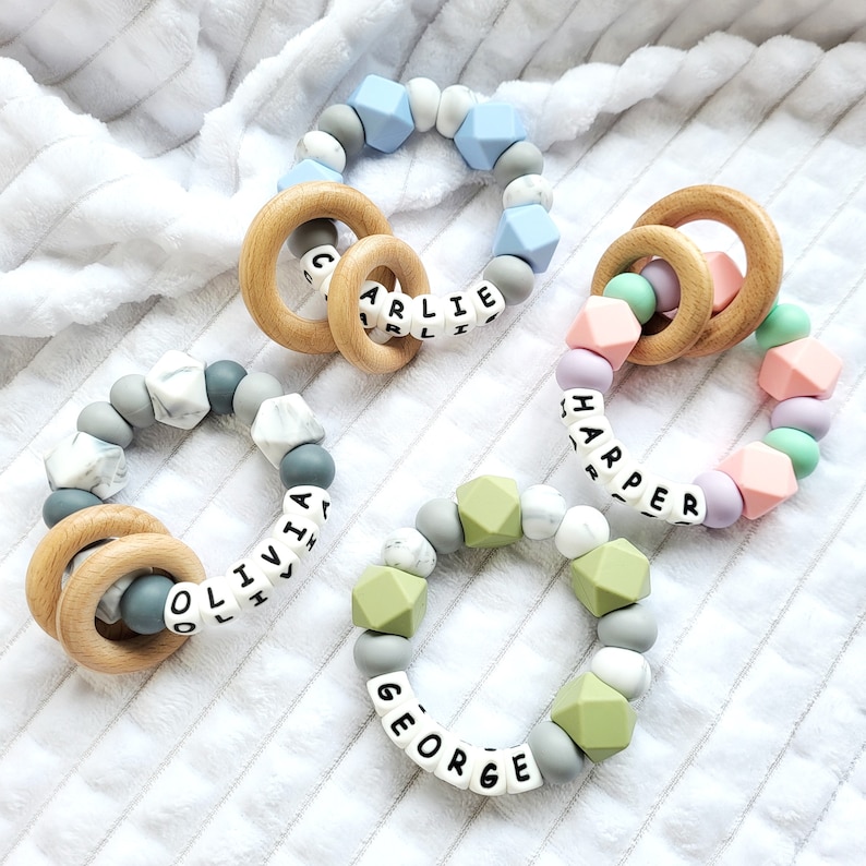 May include: Four silicone teething rings with wooden rings and personalized names: Olivia, Charlie, Harper, and George. The rings are in various colors, including gray, blue, green, and pink.
