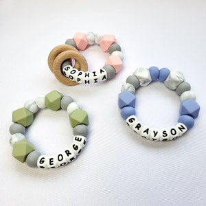 May include: Three silicone teething rings with different color beads and personalized names: Sophia, George, and Grayson. The rings are made with a combination of marble-patterned, solid color, and geometric beads.