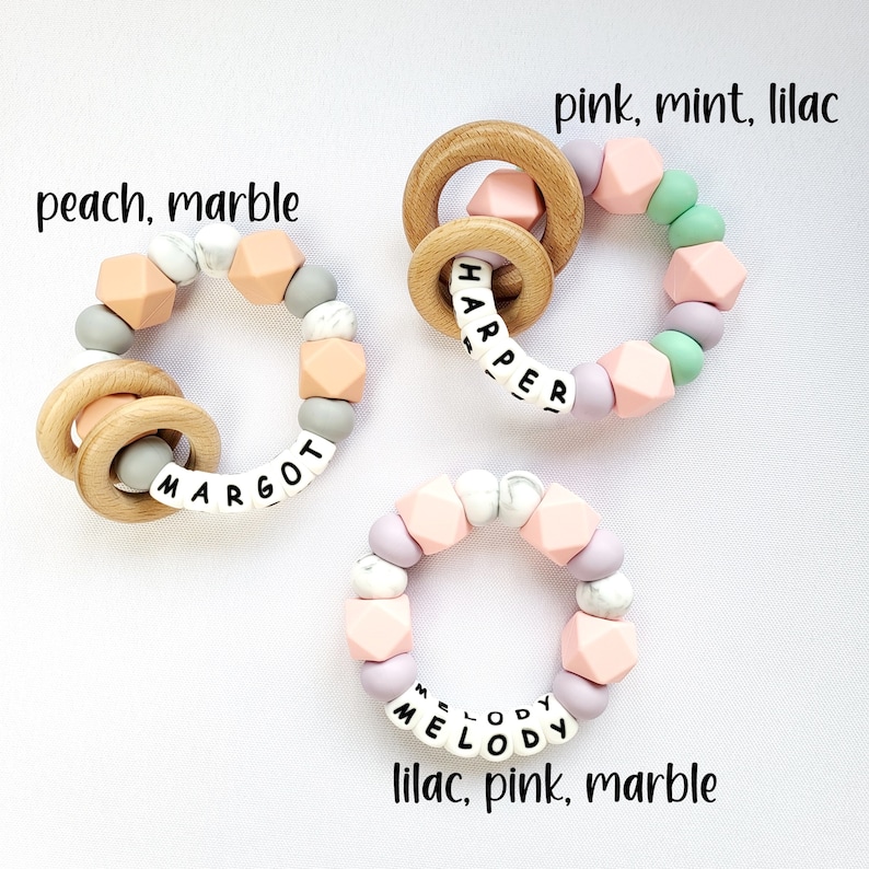 May include: Three silicone teething rings with wooden rings, each with a different name spelled out in white letters. The rings are in peach, marble, and pink, mint, lilac color combinations.