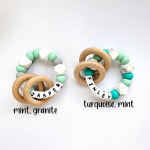 May include: Two wooden teething rings with silicone beads. One ring has mint green and white beads with the name "Jasper" spelled out. The other ring has turquoise and white beads with the name "Finley" spelled out.