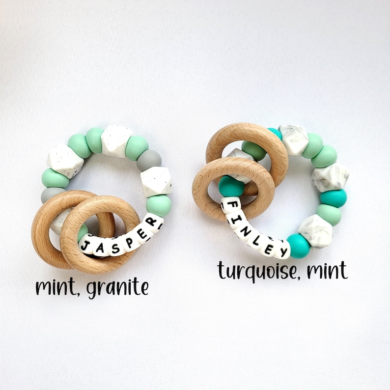May include: Two wooden teething rings with silicone beads. One ring has mint green and white beads with the name "Jasper" spelled out. The other ring has turquoise and white beads with the name "Finley" spelled out.