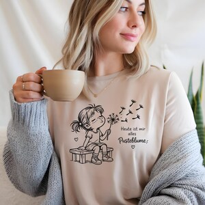 Puede incluir: Una camiseta de color beige con un gr&aacute;fico de l&iacute;neas negras de un ni&ntilde;o sentado en un banco soplando un diente de le&oacute;n. El texto dice "Heute ist mir alles Pusteblume", acompa&ntilde;ado de peque&ntilde;os iconos de corazones y semillas volando.