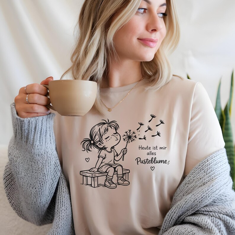 Puede incluir: Una camiseta de color beige con un gr&aacute;fico de l&iacute;neas negras de un ni&ntilde;o sentado en un banco soplando un diente de le&oacute;n. El texto dice "Heute ist mir alles Pusteblume", acompa&ntilde;ado de peque&ntilde;os iconos de corazones y semillas volando.