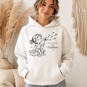 Puede incluir: Una sudadera con capucha blanca que presenta un gr&aacute;fico de l&iacute;neas negras de un ni&ntilde;o sentado en un banco soplando un diente de le&oacute;n. El texto dice: Heute ist mir alles Pusteblume. El dise&ntilde;o incluye peque&ntilde;os corazones y semillas volando.