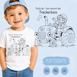 Puede incluir: Una camiseta blanca con un gráfico de líneas negras de un niño conduciendo un tractor con el texto en alemán 'Platz da - hier kommt der Treckerboss'. El diseño incluye un detalle de corazón y un parche en el capó del tractor.