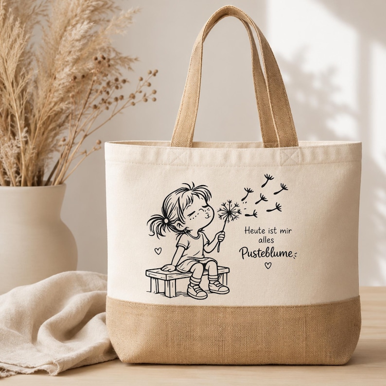 Puede incluir: Un bolso de tela color crema con una base de arpillera tostada y dos asas a juego. El frente presenta un dibujo lineal negro de un ni&ntilde;o sentado en un banco soplando un diente de le&oacute;n, con el texto en alem&aacute;n "Heute ist mir alles Pusteblume".