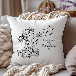 Puede incluir: Un coj&iacute;n cuadrado blanco con una ilustraci&oacute;n de l&iacute;neas negras de un ni&ntilde;o sentado en un banco soplando un diente de le&oacute;n. El texto dice "Heute ist mir alles Pusteblume" junto con peque&ntilde;os iconos de corazones y semillas volando.