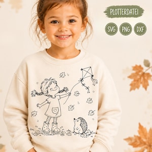 Op de afbeelding: Een crèmekleurig sweatshirt met een zwarte lijntekening van een kind met vlechten in een jurk en laarzen, staand in een plas terwijl het een vlieger vasthoudt. Een kleine egel staat ernaast, omringd door vallende herfstbladeren.