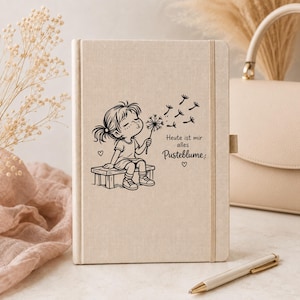 Puede incluir: Un cuaderno con cubierta de tela beige que presenta un dibujo lineal negro de un ni&ntilde;o sentado en un banco soplando un diente de le&oacute;n. El texto dice: Heute ist mir alles Pusteblume. El cuaderno incluye una banda el&aacute;stica vertical de cierre.